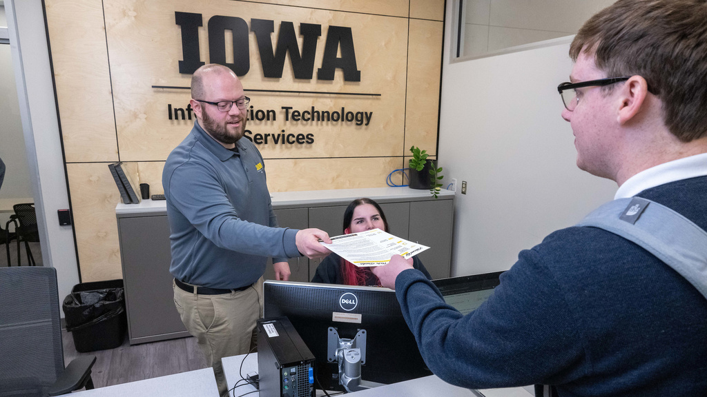 An ITS employee hands a paper to a person. Another ITS employee sits at a computer.